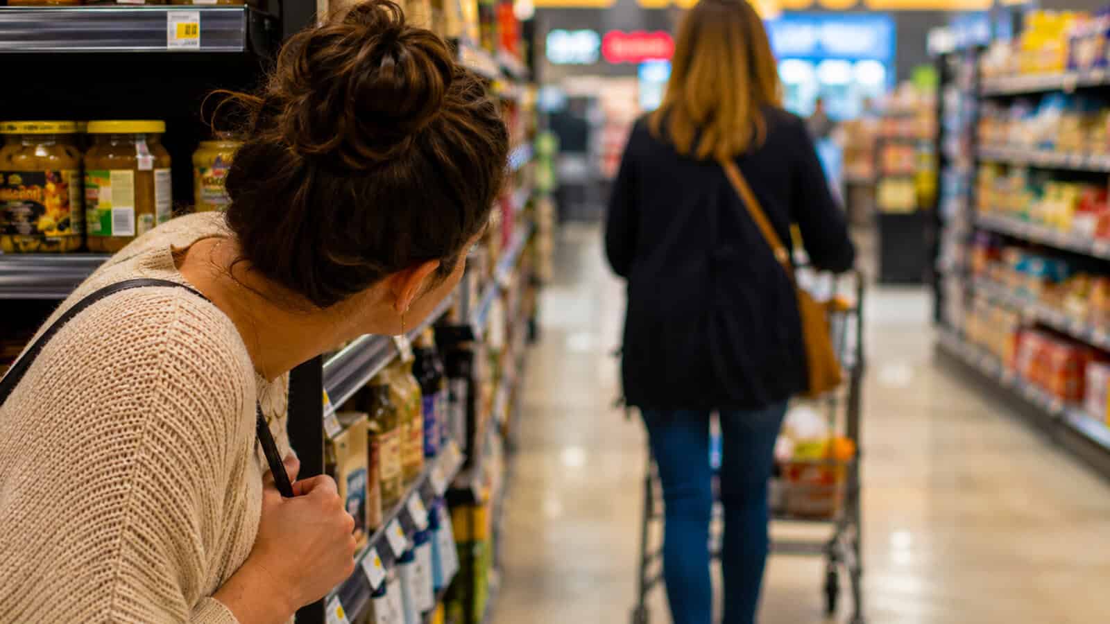 A woman peeks around a grocery store shelf, curious about the psychology behind an unplanned conversation as she watches another woman with a shopping cart walking down the aisle.