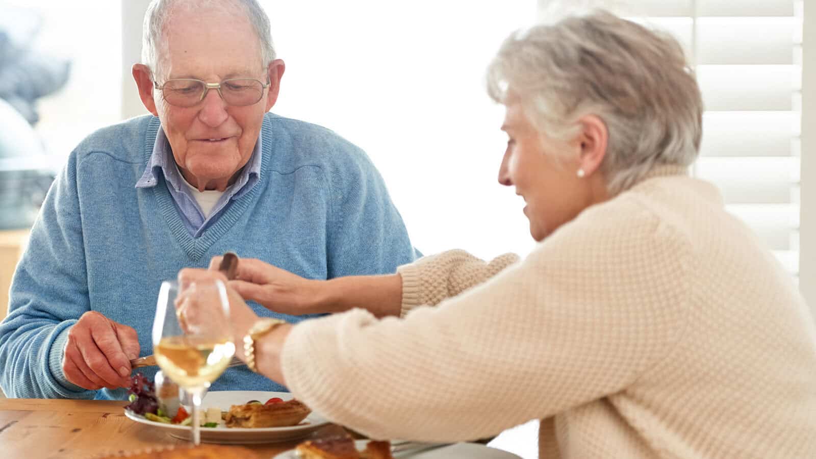 An elderly couple sits at a table enjoying an early dinner together, reflecting the psychology of shared routines and mutual approval. The man wears glasses and a blue sweater, while the woman, in a beige sweater, smiles as she reaches across with a wine glass. Bright light streams through window blinds.