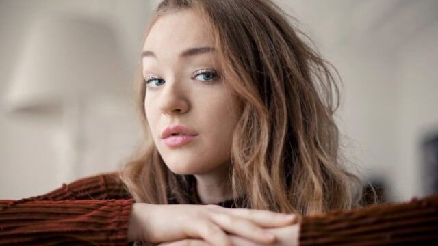 A young woman with long, wavy brown hair rests her chin on her crossed arms, gazing thoughtfully at the camera. She wears a brown, ribbed sweater and sits in a softly lit, neutral-toned room—a tranquil setting that perfectly captures the quiet contentment many introverts find in staying home.