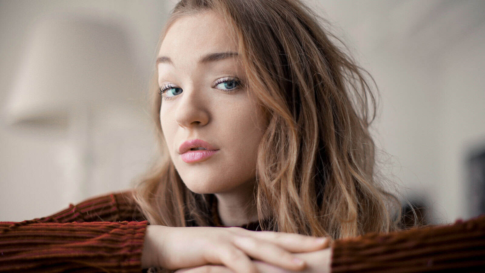 A young woman with long, wavy brown hair rests her chin on her crossed arms, gazing thoughtfully at the camera. She wears a brown, ribbed sweater and sits in a softly lit, neutral-toned room—a tranquil setting that perfectly captures the quiet contentment many introverts find in staying home.