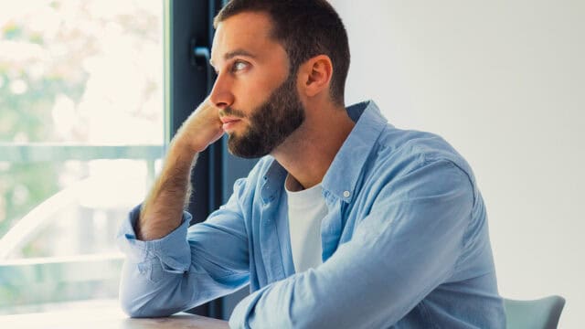 A man with a beard, wearing a light blue shirt, sits at a table and looks thoughtfully out of a window with his hand resting on his cheek, as if lost in the kind of reflection associated with psychology’s concept of the default mode network.