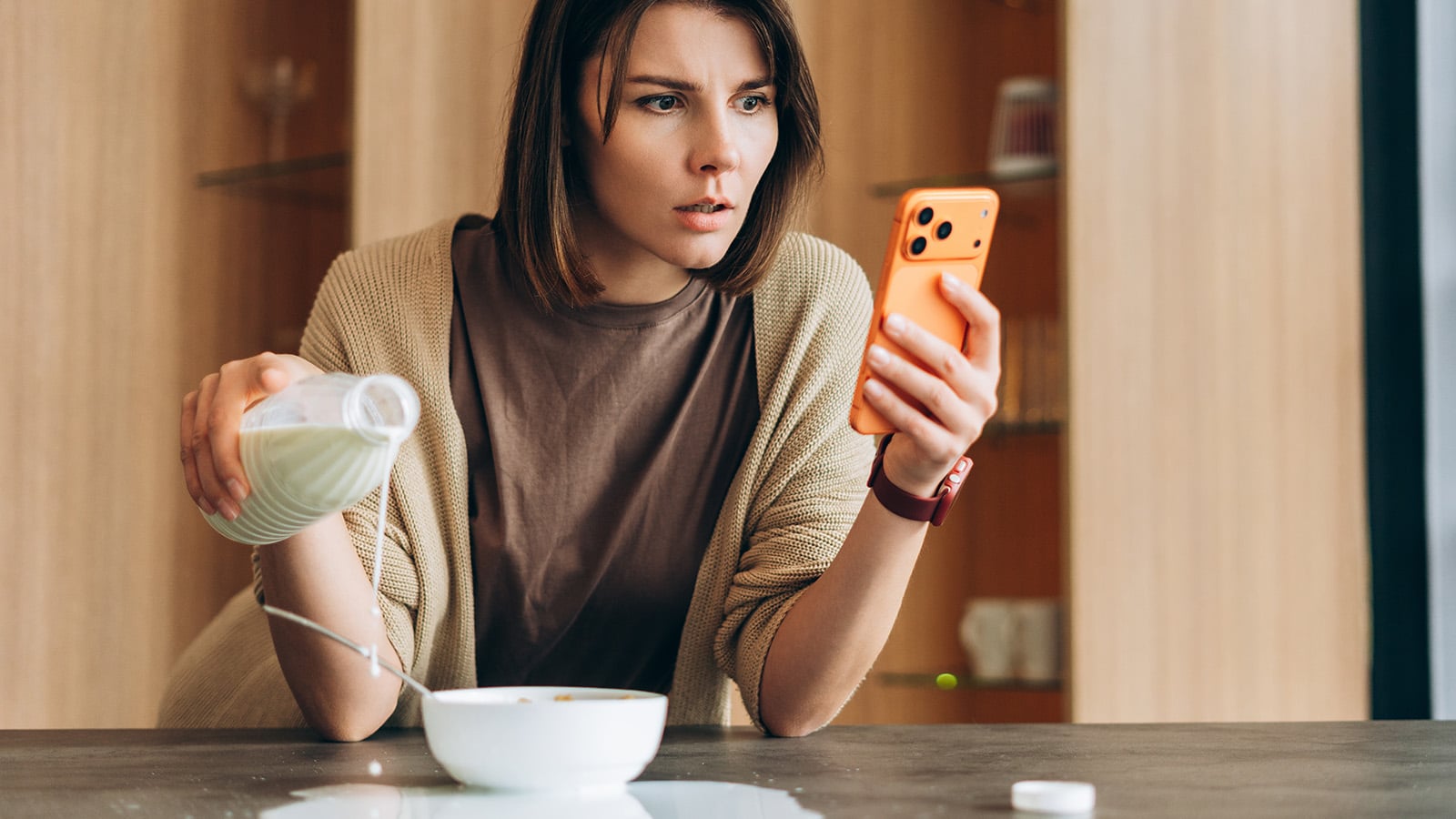 A woman with ADHD looks concerned at her smartphone, accidentally pouring milk over the edge of a bowl—highlighting how distractions can impact even simple tasks despite her intuitive reasoning.
