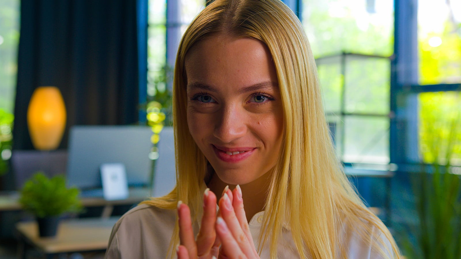 A young woman with long blonde hair smiles gently, holding her hands clasped near her face. She is indoors with natural light and greenery in the background, creating a bright and cozy atmosphere—her warm expression exuding behaviors that look generous, reflecting kindness often discussed in psychology.