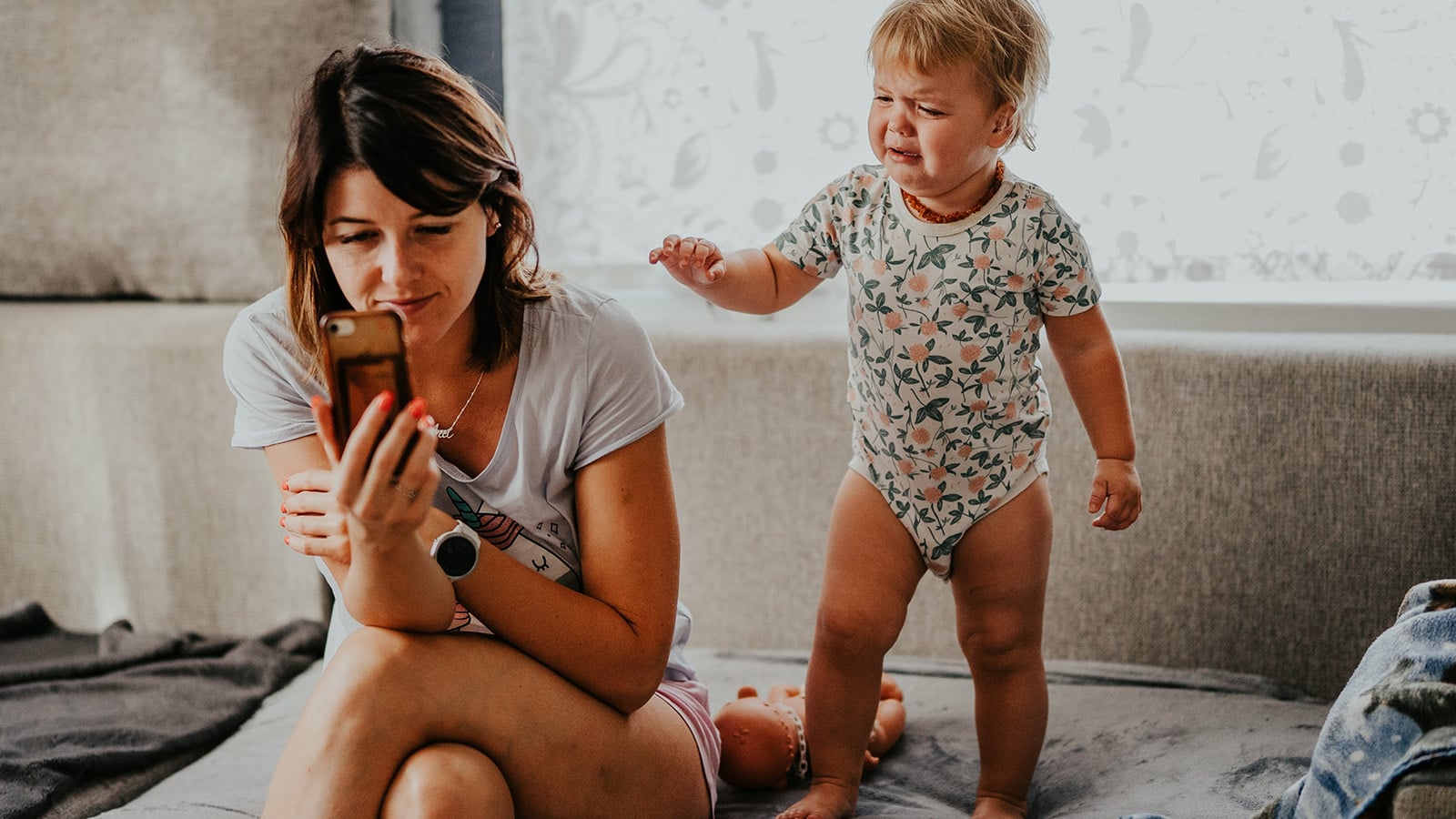 A woman sits on a couch looking at her phone while a crying toddler in a patterned onesie stands next to her, reaching for attention—a scene reminiscent of the still-faced experiment in baby psychology that highlights how parent phone use can impact child emotional responses.