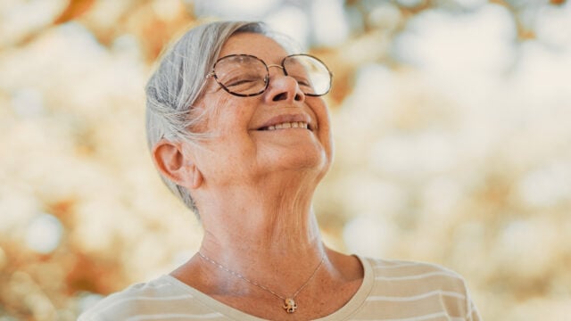 An older woman with glasses smiles peacefully with her eyes closed outdoors. She wears a striped shirt and a necklace, embodying the calm that often comes after retirement adjustment. Sunlight and blurry autumn leaves in the background create a warm setting, highlighting the anchors of comfort and tranquility that many retirees find in this new chapter of life.