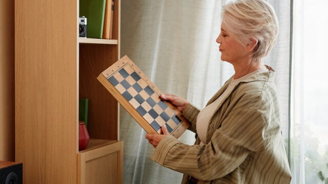 An older woman with short gray hair holds a chessboard and looks at it thoughtfully, perhaps contemplating its object value and the endowment effect, while standing next to a wooden shelf lined with books, a red vase, and a vintage camera.