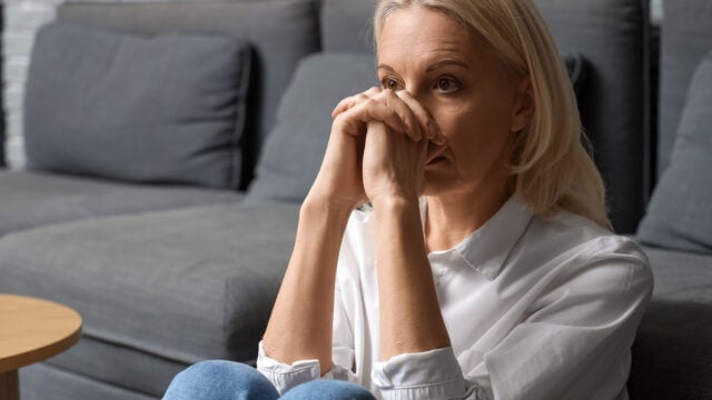 A woman with blonde hair sits on the floor in front of a gray couch, resting her chin on her hands and looking thoughtful or worried, as if carrying grief alone after heavy losses. She wears a white shirt and blue jeans.