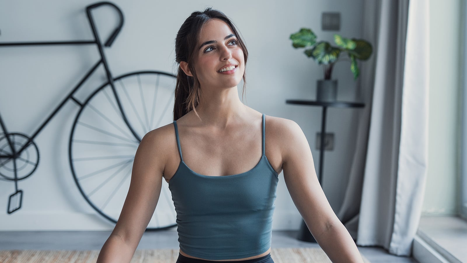 A young woman in workout clothes sits indoors, smiling and looking up—her peaceful glow hints at effective stress recovery. A modern bike decor is mounted on the wall, and a potted plant sits on a small table near a sunlit window.