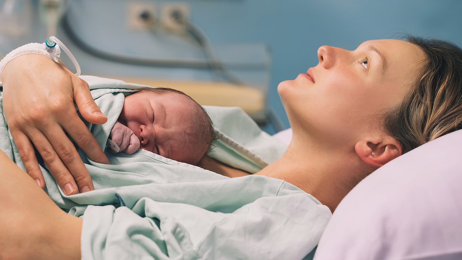 A woman lies in a hospital bed holding a newborn baby on her chest, both wrapped in hospital gowns. The woman looks upward, appearing calm and reflective, capturing the matrescence experienced by many new mothers in this hospital setting.