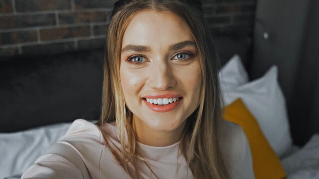 A woman with long light brown hair smiles at the camera while sitting on a bed with white linens and a yellow pillow, in a room with a dark headboard and brick wall—showing subtle signs of being the responsible eldest daughter.