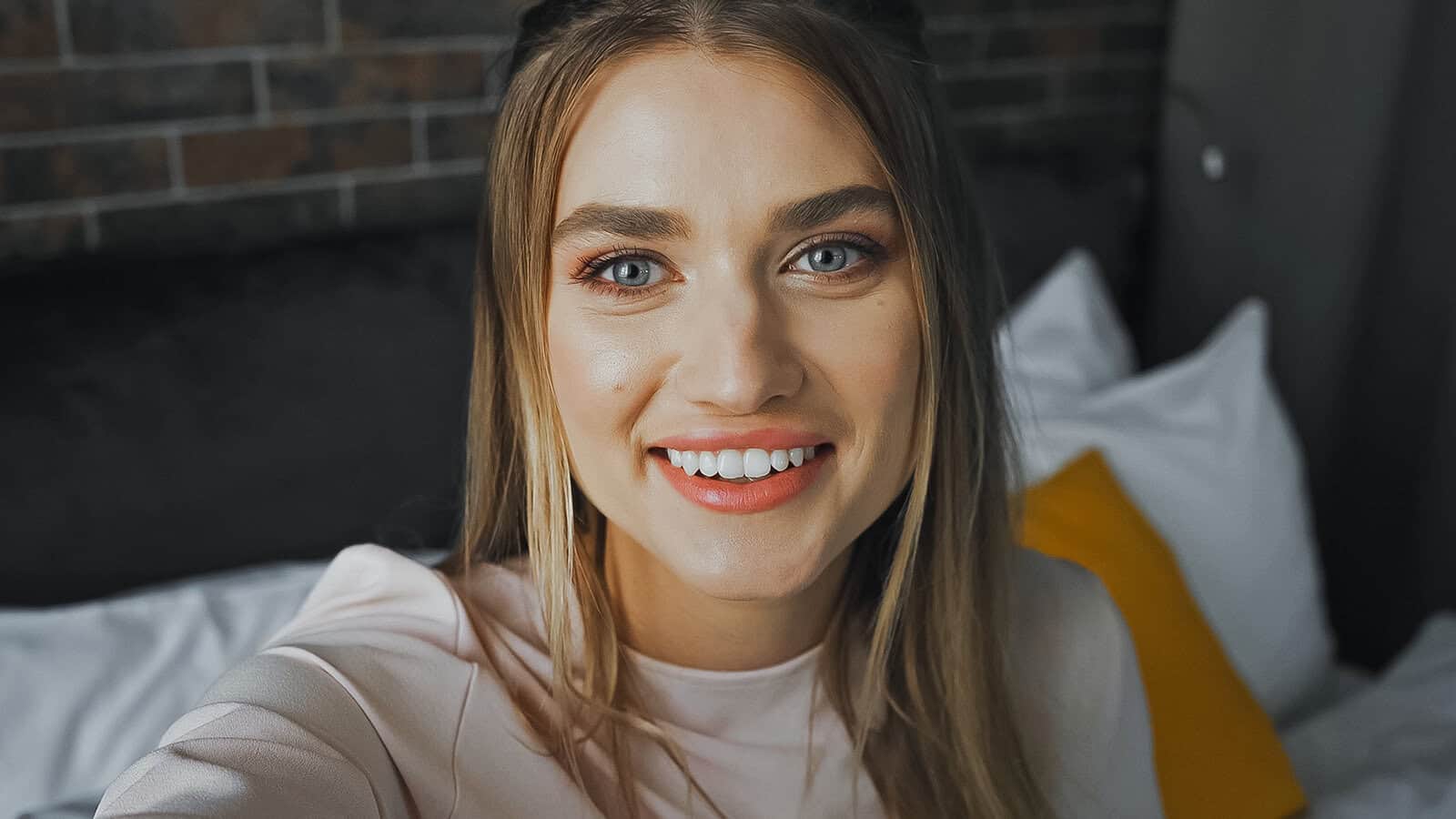 A woman with long light brown hair smiles at the camera while sitting on a bed with white linens and a yellow pillow, in a room with a dark headboard and brick wall—showing subtle signs of being the responsible eldest daughter.