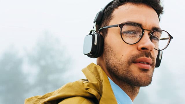 A man wearing glasses and over-ear headphones looks into the distance, as if deep in casual conversation. He has a short beard and is dressed in a yellow jacket, with a blurred outdoor background and misty trees behind him, giving an impression that he might be pondering something smarter than they actually are.