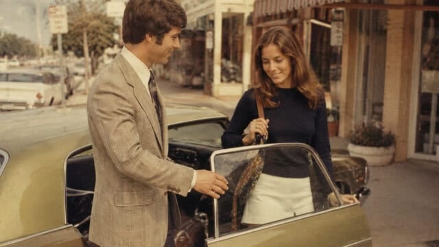 A man in a plaid suit holds a car door open for a smiling woman in a navy top and white skirt on a sunny street lined with shops.