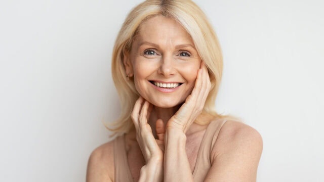 Smiling older woman with long blonde hair and blue eyes, touching her face gently with both hands. She is wearing a sleeveless beige top and stands against a plain white background.