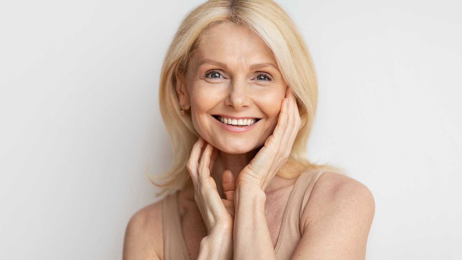 Smiling older woman with long blonde hair and blue eyes, touching her face gently with both hands. She is wearing a sleeveless beige top and stands against a plain white background.