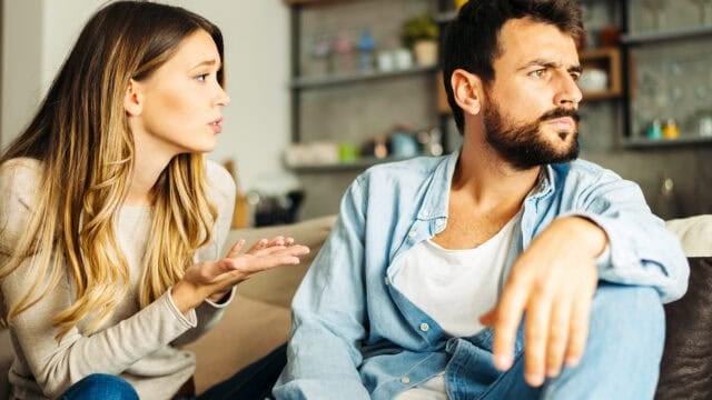 A woman appears to be talking emotionally to a man who is looking away with a serious expression while sitting on a couch in a living room.