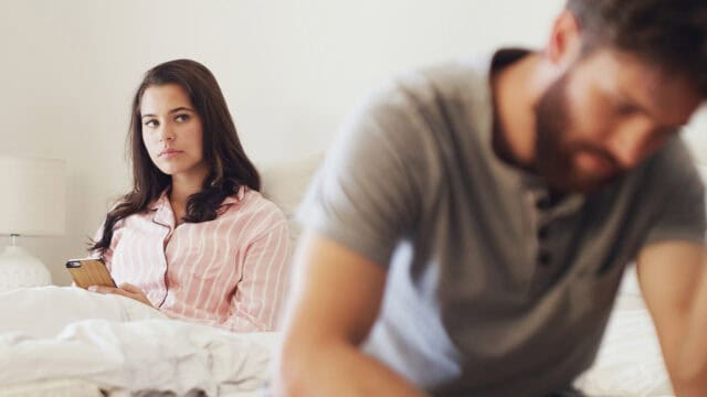 A woman in pink pajamas sits on a bed holding a phone, looking at her partner—a man in a gray shirt who sits at the edge of the bed, facing away and appearing upset—revealing subtle signs that not everything is well between them or that one may be more emotionally attached than the other.