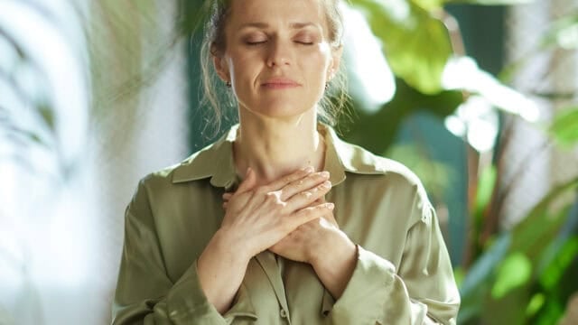 A woman with closed eyes gently places her hands over her chest, appearing calm and peaceful, standing among green plants in soft natural light.