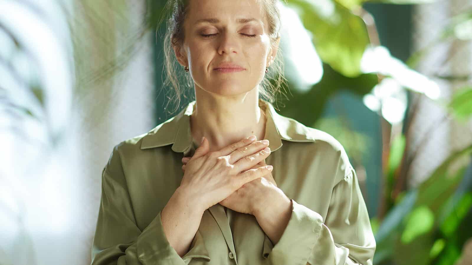 A woman with closed eyes gently places her hands over her chest, appearing calm and peaceful, standing among green plants in soft natural light.