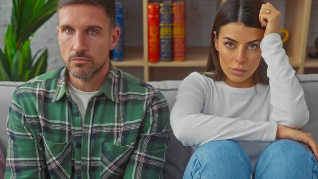 A man and a woman sit side by side on a couch, both looking serious and slightly tense. Shelves with books and a plant are in the background.
