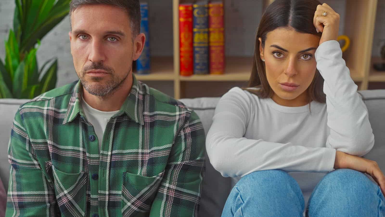 A man and a woman sit side by side on a couch, both looking serious and slightly tense. Shelves with books and a plant are in the background.