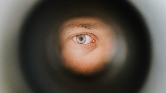 A close-up of a blue eye peering through a circular opening, with the surrounding area blurred to create a tunnel-like effect.