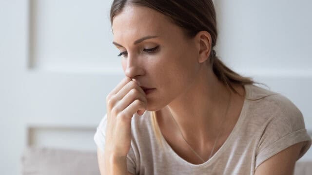 A young woman with brown hair in a ponytail sits indoors, resting her hand near her mouth and looking down thoughtfully, appearing worried or deep in thought.