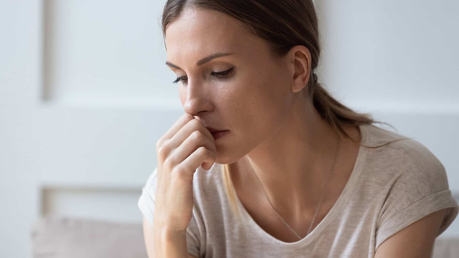 A young woman with brown hair in a ponytail sits indoors, resting her hand near her mouth and looking down thoughtfully, appearing worried or deep in thought.