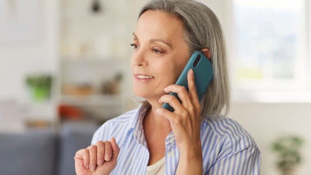 Smiling older woman with gray hair and a striped shirt holds a teal smartphone to her ear, appearing to enjoy a pleasant phone conversation in a bright, cozy room—showcasing the strong social skills often seen in people in their 50s and 60s.