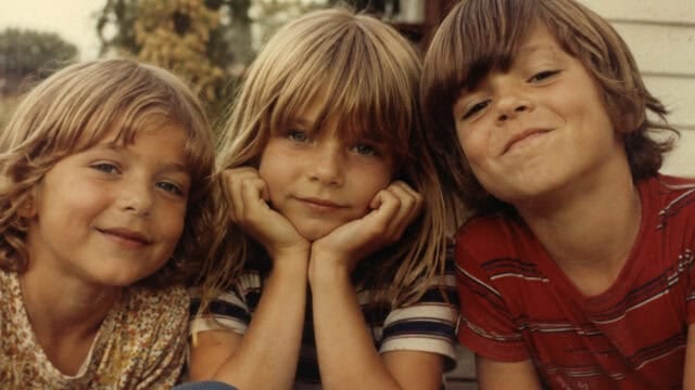 Three young children with light brown hair sit close together outside, smiling at the camera. The child in the center rests their chin on their hands, while the others lean in from each side.