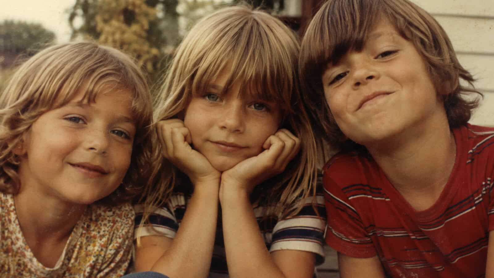 Three young children with light brown hair sit close together outside, smiling at the camera. The child in the center rests their chin on their hands, while the others lean in from each side.
