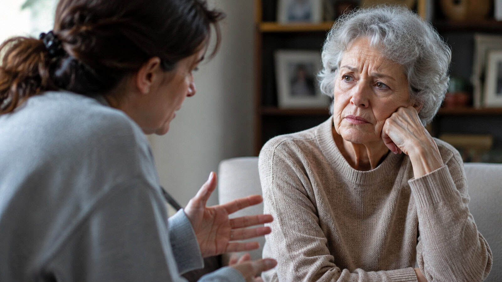 An older woman listens thoughtfully with her hand on her chin while another person—perhaps one of her adult children—speaks to her, gesturing with their hands. They appear to be having a serious conversation in a cozy indoor setting, possibly discussing whether parents over 70 should consider downsizing.