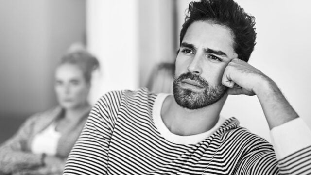 A man with a beard in a striped shirt rests his chin on his hand, looking thoughtful. In the blurred background, a woman appears to be sitting and looking in his direction. The image is in black and white.