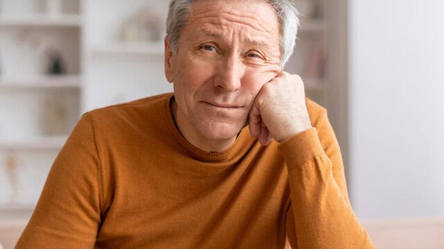 An older man in an orange sweater rests his head on his hand, looking thoughtfully at the camera. The background is softly focused, showing light-colored shelves.