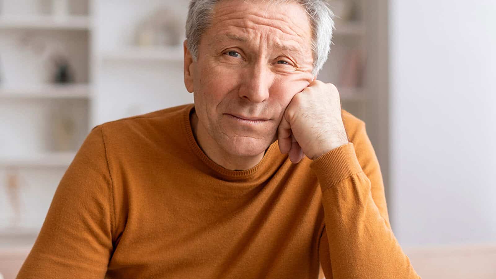 An older man in an orange sweater rests his head on his hand, looking thoughtfully at the camera. The background is softly focused, showing light-colored shelves.
