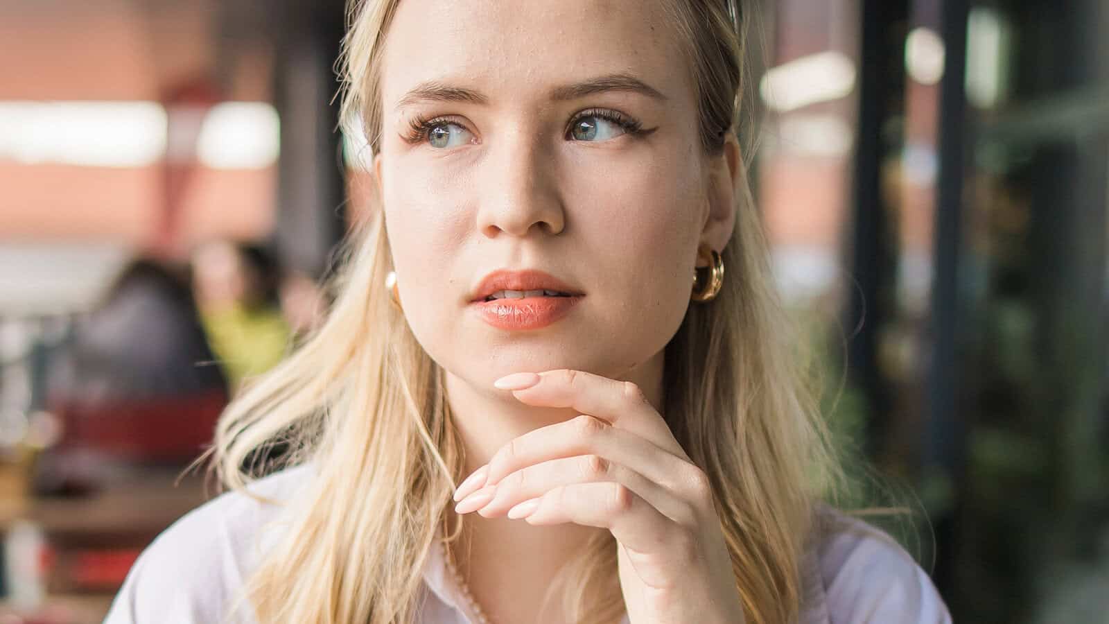 A blonde woman with light eyes sits indoors, looking thoughtfully to the side with her hand near her chin—a quiet moment characteristic of observant people. She wears gold hoop earrings and a light top, with a softly blurred background.