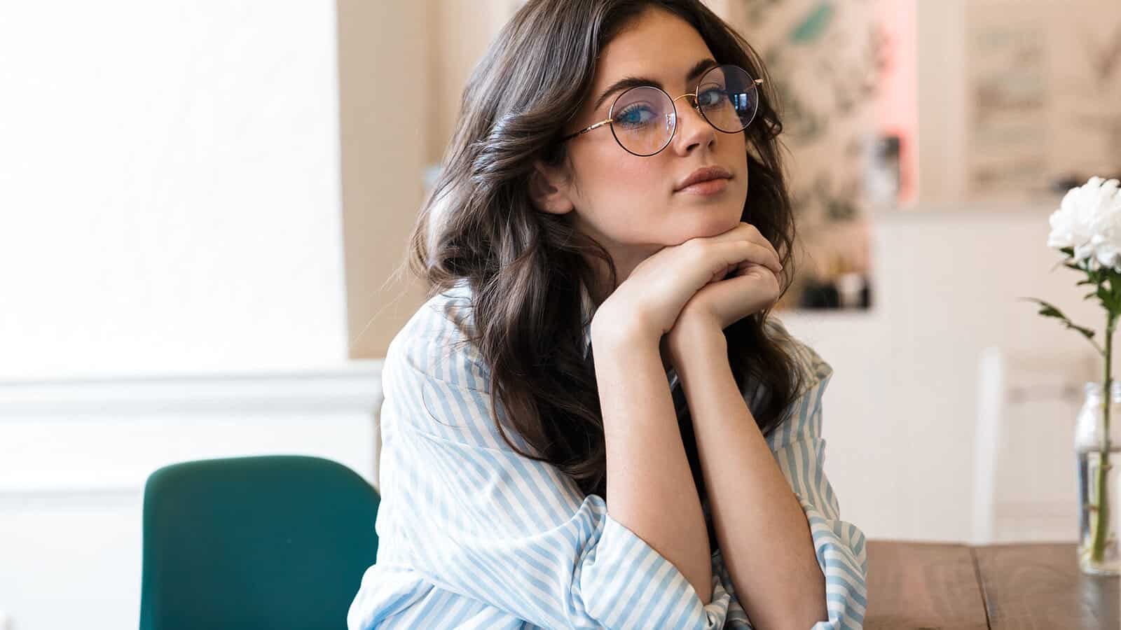 A young woman with long brown hair and round glasses sits indoors at a table, resting her chin on her hands and looking thoughtfully ahead. She wears a light blue striped shirt. A white flower is on the table beside her.