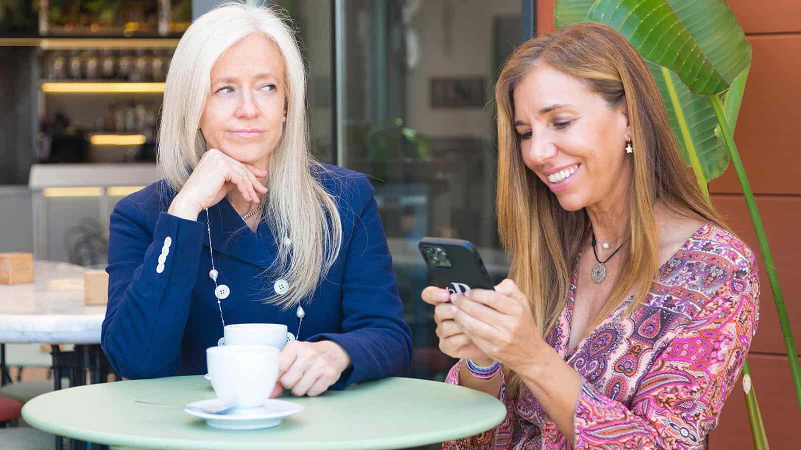 Two women sit at a cafe table. One woman looks annoyed—clear signs of outgrowing a friendship—while the other smiles at her phone, seemingly oblivious. There are coffee cups on the table and a green plant in the background.