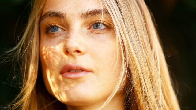 A close-up of a young person with long blonde hair and blue eyes, looking slightly away. Sunlight creates subtle shadows and highlights on their face, with strands of hair falling gently across their features.