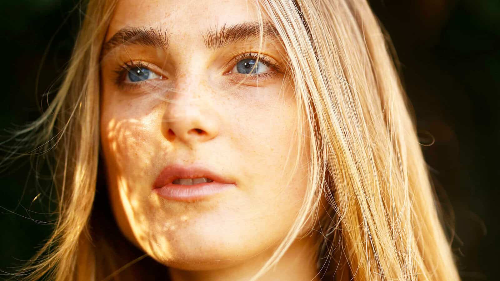 A close-up of a young person with long blonde hair and blue eyes, looking slightly away. Sunlight creates subtle shadows and highlights on their face, with strands of hair falling gently across their features.