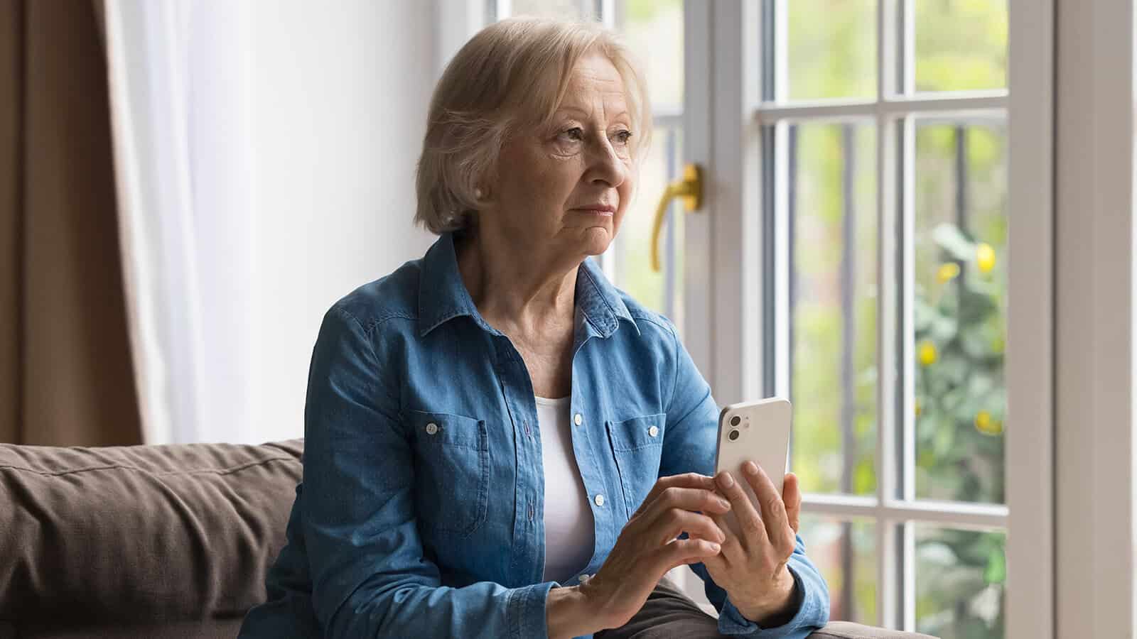 An older woman with gray hair sits on a couch by a window, holding a smartphone and looking thoughtfully outside.