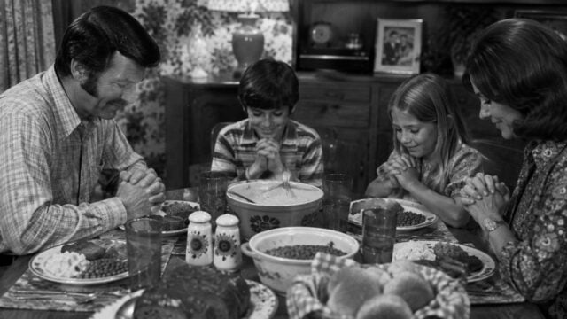 A family of four sits at a dinner table with various dishes, heads bowed and hands clasped in prayer, sharing a moment of gratitude before their meal.