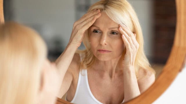 A middle-aged woman with blonde hair looks at herself in a mirror, gently touching the skin on her forehead with both hands, appearing thoughtful.
