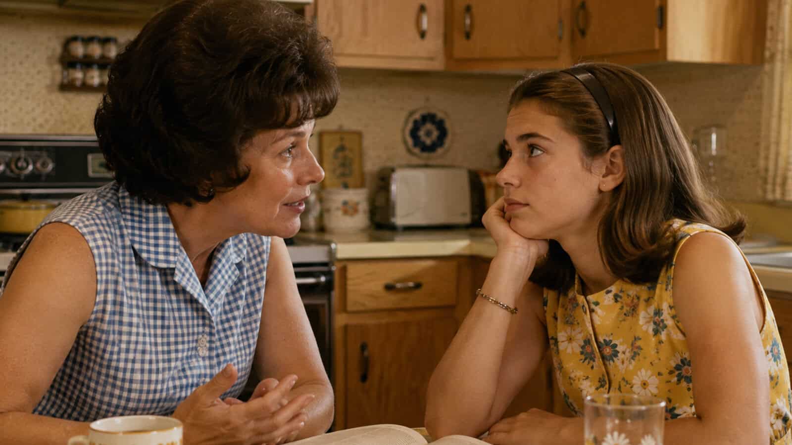 A woman in a blue checkered blouse talks seriously to her daughter in a yellow dress, who listens with her chin in her hand at a kitchen table. The kitchen is decorated in a vintage style, evoking the warmth and wisdom often shared between boomer mothers and their daughters.