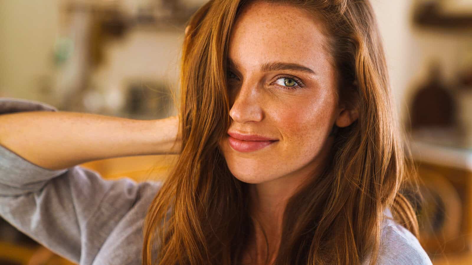 A woman with long auburn hair and freckles smiles softly at the camera, her hand touching her hair. She has green eyes and is wearing a light gray shirt in a warmly lit indoor setting, radiating a quiet self reliance that suggests master-level confidence.