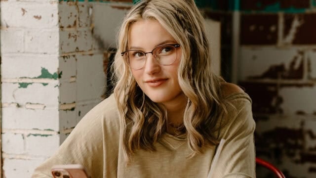A young woman with wavy blonde hair and glasses smiles slightly while sitting indoors, holding a phone. She wears a beige top and sits in front of a white brick wall.