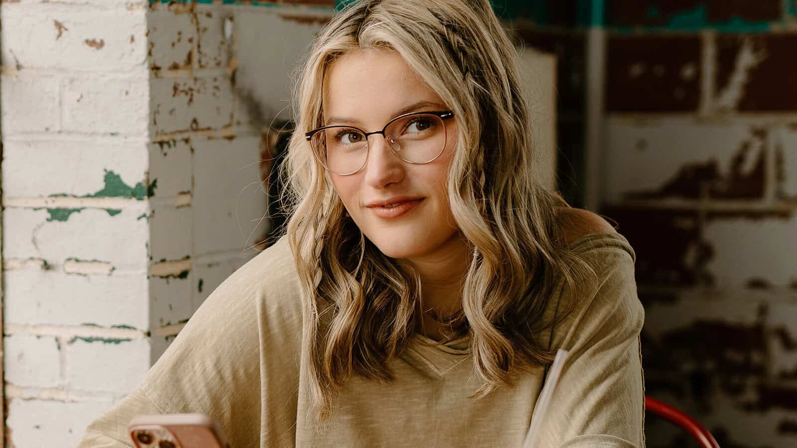 A young woman with wavy blonde hair and glasses smiles slightly while sitting indoors, holding a phone. She wears a beige top and sits in front of a white brick wall.