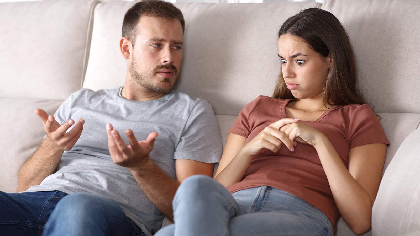 A man and woman sit on a couch. The man gestures with his hands, appearing frustrated, while the woman looks away with a surprised expression and her fingers intertwined.