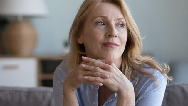 A middle-aged woman with blonde hair rests her chin on her hands, looking to the side and smiling softly. She is sitting indoors on a couch, wearing a light blue shirt. The background is softly blurred.