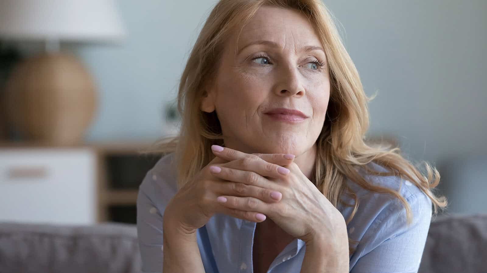A middle-aged woman with blonde hair rests her chin on her hands, looking to the side and smiling softly. She is sitting indoors on a couch, wearing a light blue shirt. The background is softly blurred.